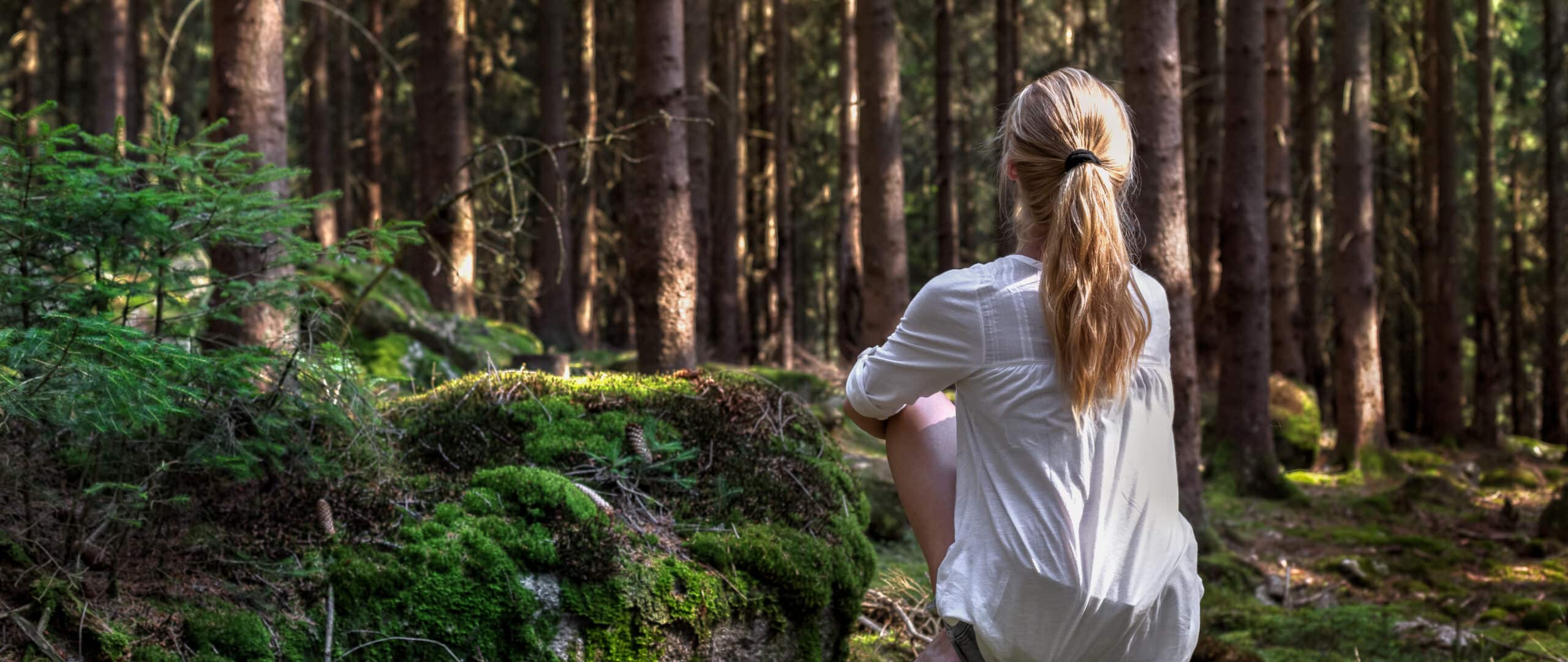 Kiven päällä metsässä istuu nainen kasvot pois päin